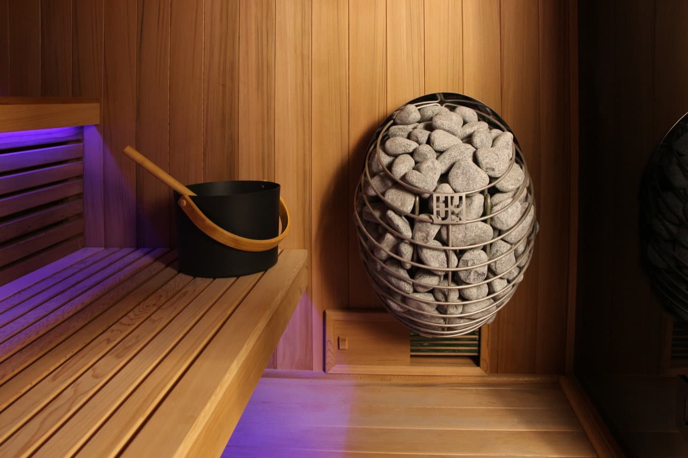HUUM Drop heater inside the Nova: a mesh basket of stones mounted on the cedar wall above the vent, with a bucket and ladle on the bench
